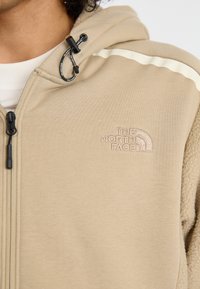 Sudadera beige que presenta una textura de felpa por un lado, tela suave por el otro, cordón ajustable negro y logo bordado en el pecho.