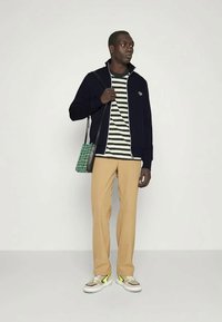 Jūras spēku zippers jakas, svītrainā zaļā un baltā krekliņa, bēša bikses un sporta apavi ar dzelteniem akcentiem; modele tur zaļu rūtotu somu.