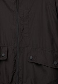 Tissu de veste noire avec fermeture éclair centrale, deux poches frontales à rabat sécurisées par des boutons-pression noirs ronds, et détail de poche cousu verticalement au-dessus de la fermeture éclair.