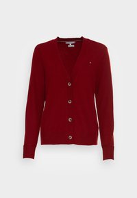 Červený svetr na zapínání z měkké tkaniny s hlubokým výstřihem do V, dlouhými rukávy, šesti hnědými knoflíky vpředu a malým logem na hrudi.