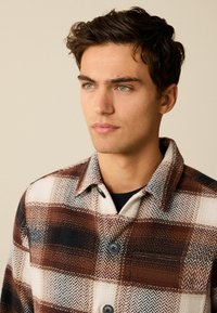 Brun og creme ternet skjortejakke med et houndstooth-mønster, to sorte knapper, en krave og en blød tekstur.