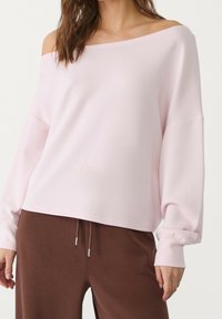 Hellrosa schulterfreier Pullover mit weiten Ärmeln und gerippten Bündchen, kombiniert mit braunen Hosen mit Kordelzug. Weiche, glatte Stofftextur.