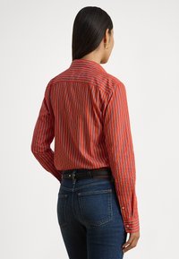 Červeno-bielo pruhovaná košeľa na gombíky s golierom, s dlhými rukávmi a prispôsobeným strihom, nosená s tmavomodrými džínsami.