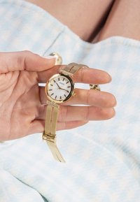 Montre-bracelet avec un bracelet en mailles dorées et un cadran blanc rond, présentant des marqueurs des heures dorés et une couronne, tenue à la main contre un fond à motifs.