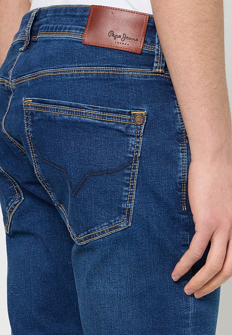 Schmal geschnittene blaue Jeans aus Denim mit einem braunen Lederrückenpatch, goldenen Nähten und einer hinteren Tasche mit einem runden Metallnieten.