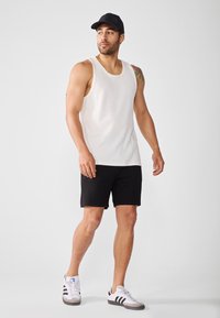 En mand står i en hvid ribbet tanktop og sorte shorts, iført en sort kasket og hvide sneakers, poserende mod en lys baggrund.