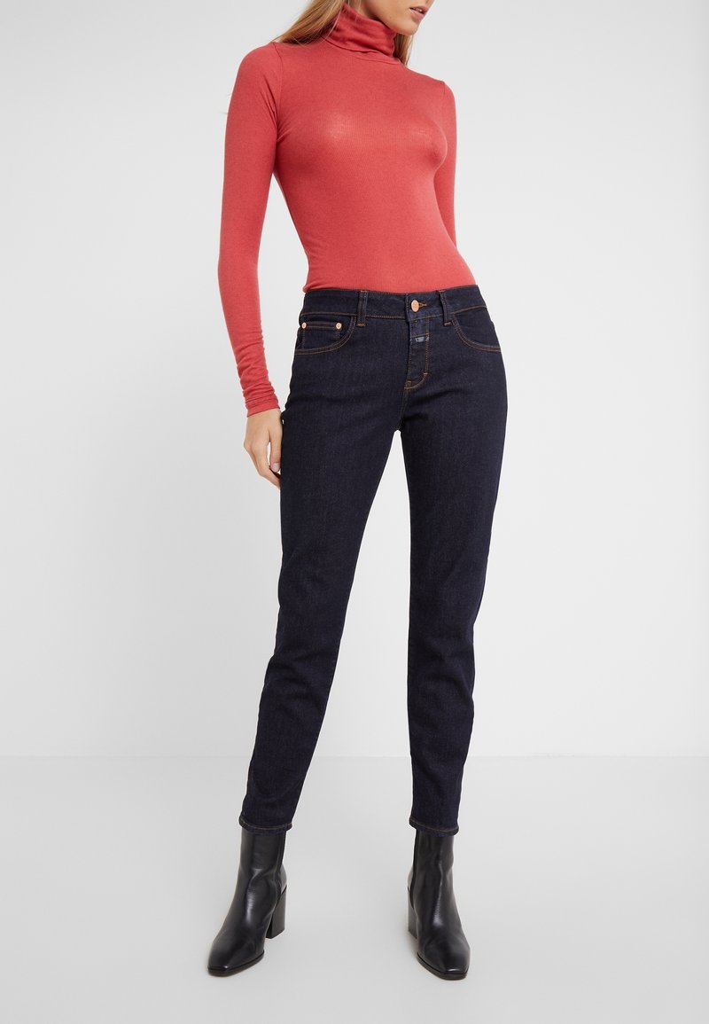 Červený body s rolákem, kombinovaný s tmavě modrými skinny džínami. Černé kotníkové boty doplňují tento outfit. Čistý, minimalistický design a hladká textura.