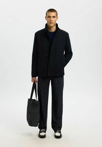 Cappotto in misto lana blu navy con chiusura a bottoni, colletto rialzato, abbinato a pantaloni scuri con texture e una borsa tote in tessuto nera.