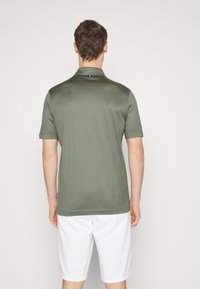 Olivgrön kortärmad polo med en slät yta, som har en krage och en liten märkesskylt vid fållen.