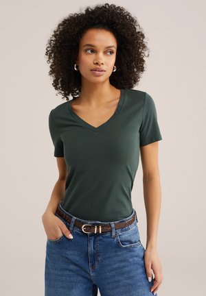 Mujer con cabello rizado, vistiendo una camiseta de cuello en V verde oscuro, jeans azules y cinturón marrón con estampado, de pie con una mano en el bolsillo.