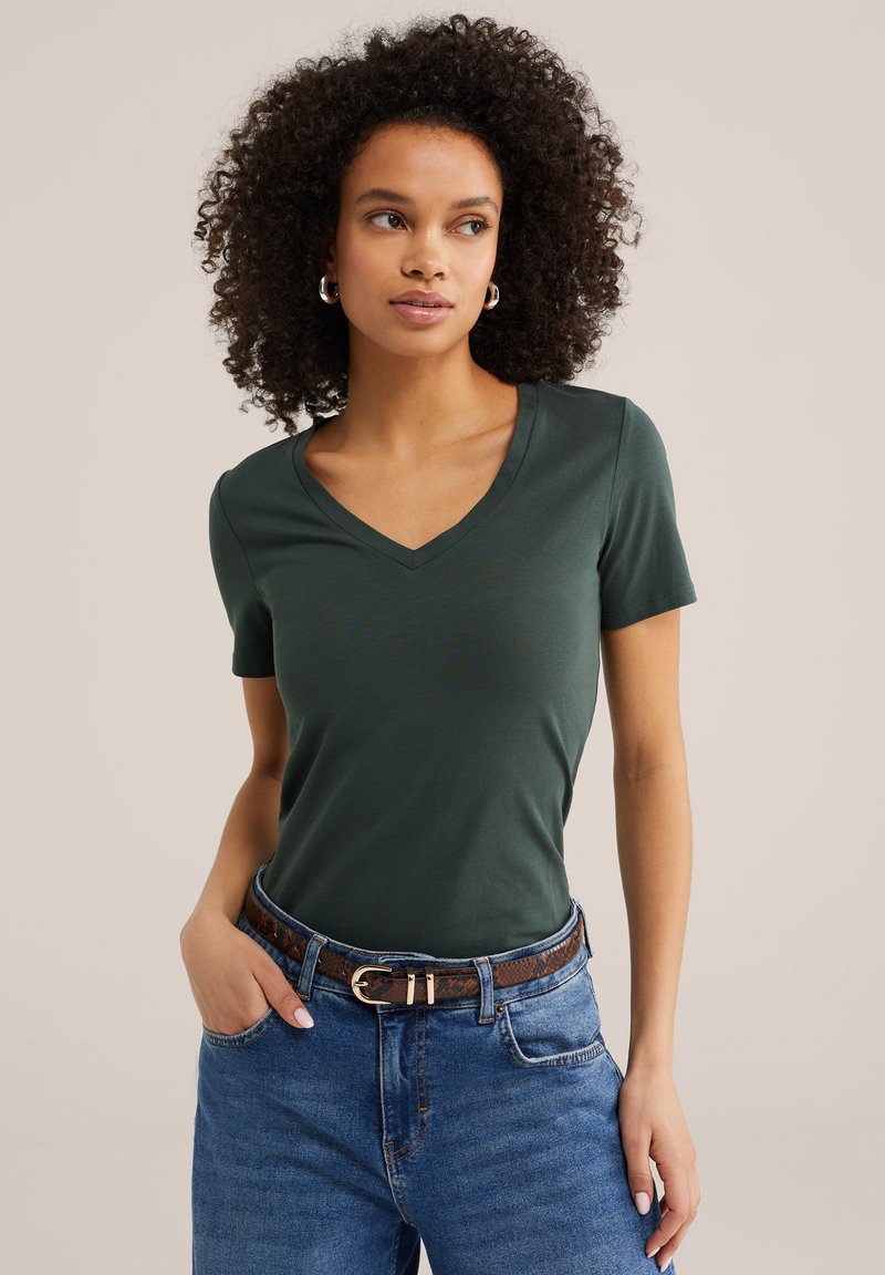 Göndör hajú nő sötétzöld V-nyakú pólóban, kék farmerben és barna mintás övben, kezét a zsebében tartva áll.