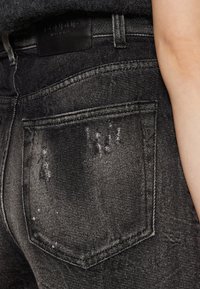 Jean en denim noir avec une texture usée, comprenant une poche arrière et un petit logo sur la ceinture.