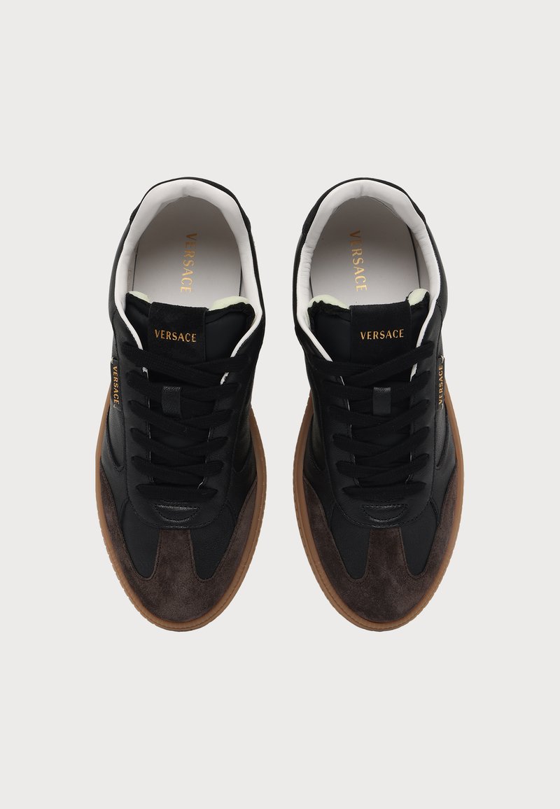 Paar schwarze und braune Versace-Sneaker mit weißem Innenfutter, schwarzen Schnürsenkeln und goldenem Versace-Branding auf der Zunge und an der Seite.