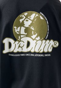 Černé tričko s velkými zlatými a bílými grafikami, včetně globu a značky "Dr. Denim", s textem níže uvádějícím původ a motto.