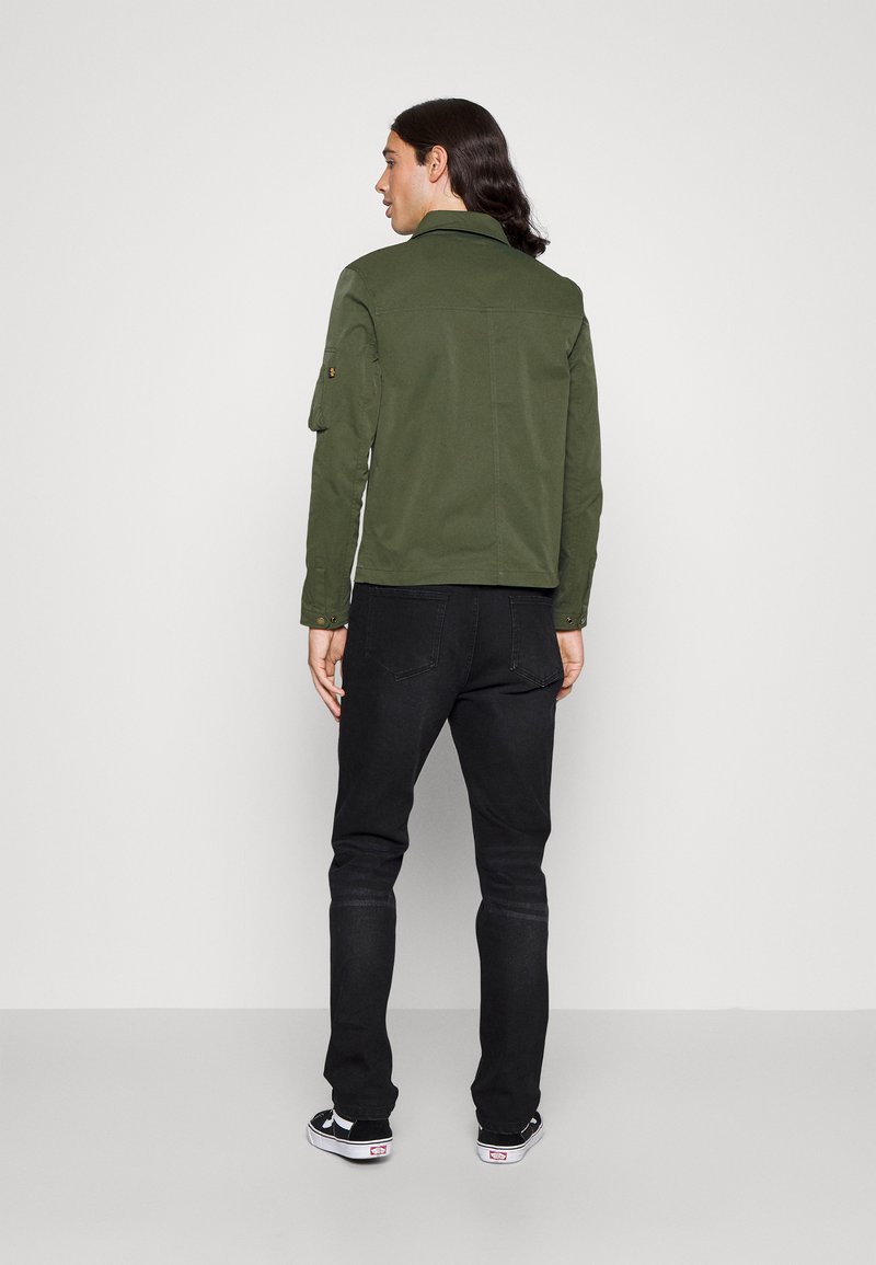 Chaqueta de lona verde con cuello doblado, mangas largas y un bolsillo en el brazo con botón, combinada con jeans negros ajustados y zapatillas negras de patrón de tablero de ajedrez.