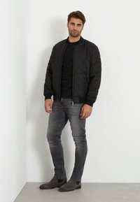 Svart quiltad bomberjacka med ribbade muddkanter och fåll, baren över en svart T-shirt. Grå slim-fit jeans och grå mocka ankelstövlar fulländar outfiten.