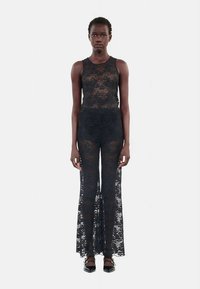 Femme portant un haut en dentelle noire sans manches et un pantalon large en dentelle assorti, se tenant droite contre un fond clair et uni.