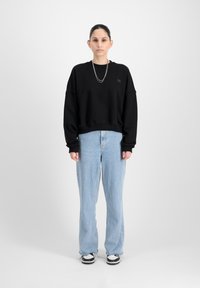 Felpa nera con spalle cadenti, dotata di un piccolo logo applicato. Jeans baggy azzurri con orlo sfrangiato e scarpe da ginnastica nere.