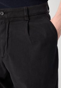 Pantalons en coton noir avec une coupe droite, fermeture à bouton sur le devant et poches latérales. Texture lisse avec des plis subtils au niveau de la taille.
