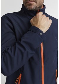 Giacca softshell navy con cerniera e dettagli arancioni; presenta un colletto alto e dettagli con bottoni a scatto sui polsini. Texture liscia, vestibilità atletica.