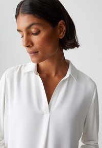 Weiße Bluse mit Kragen, weichem Stoff und V-Ausschnitt. Lange Ärmel mit glatter, untexturierter Oberfläche.