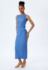 Vestido azul sin mangas con tela texturizada y detalle fruncido en la cintura, que fluye hasta una longitud de media pantorrilla. Complementado con sandalias de tacón amarillas.