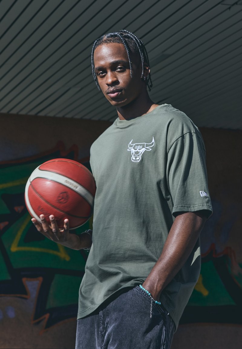 Olivenfarvet bomulds t-shirt med et hvidt okselogo, båret sammen med grå denimshorts. Personen holder en rød og hvid basketball.