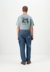 Man wearing blue graphic t-shirt and loose blue jeans with brown shoes, standing facing a plain white background.