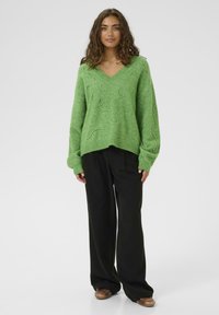 Suéter verde con cuello en v y patrones texturizados en espiral, hecho de material suave. Combinado con pantalones negros de pierna ancha y calzado marrón.