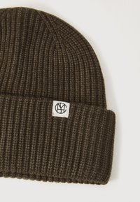 Braune gestrickte Mütze mit gerippter Textur, die ein weißes Etikett mit einem runden Logo zeigt. Cuff-Design mit enger Passform.