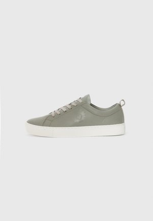 Lichtgrijze sneaker van soepel materiaal, met een witte zool, platte veters en een klein logo op de zijkant. Eenvoudig, laag profiel ontwerp.