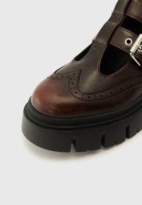 Scarpa in pelle marrone con dettagli perforati, punta arrotondata, inserti con fibbia argentata e una robusta suola in gomma nera.