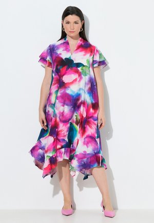 Kobieta ubrana w żywą, kwiecistą sukienkę midi z falbankowymi rękawami i dołem, w różowych szpilkach, stojąca na tle białego, gładkiego tła.