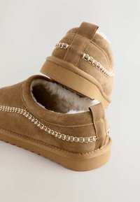 Pruunist seemisnahast slip-on kingad, millel on valge vale karv vooder, dekoratiivne tikandi detail ja tekstuuriga kummitald.