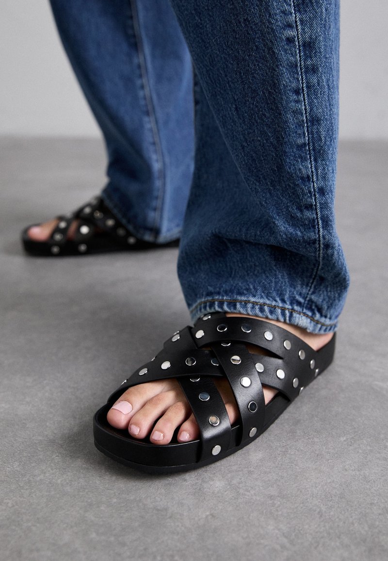 Sandales noires à semelle plate avec des brides en cuir croisées, ornées de clous en argent. Portées avec un jean en denim bleu, laissant apparaître les orteils et la couleur naturelle des ongles.