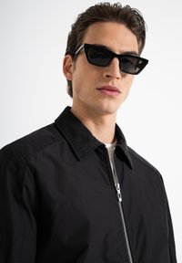 Giacca nera con zip e colletto a punta, caratterizzata da una trama liscia. Abbinata a occhiali da sole neri angolari, creando un look moderno.