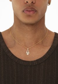 Zlatna ogrlica s privjeskom u obliku hamsa ruke ukrašena plavim detaljima, nosi se na teksturiranoj smeđoj majici s rebrastim uzorkom, a prikazana je izbliza kako bi se istaknula predmet nakita.