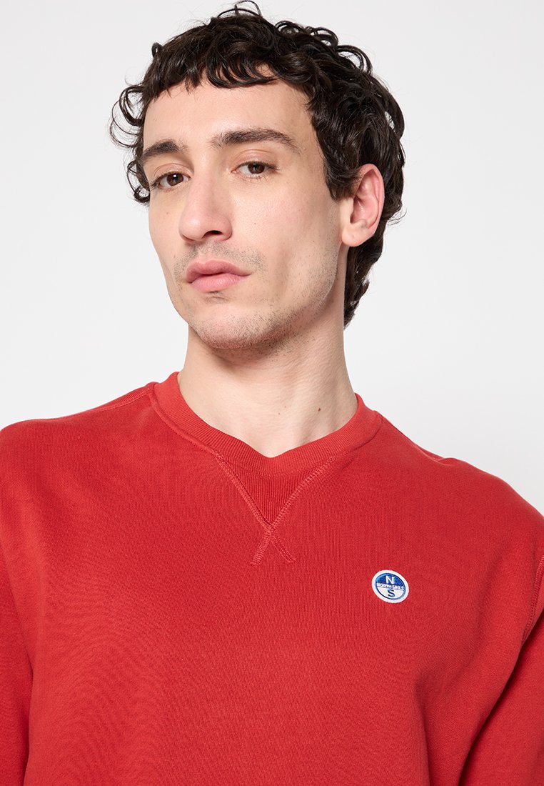 Felpa rossa in un tessuto morbido con scollatura a V. Presenta un piccolo logo circolare sul lato sinistro del petto e dettagli della cucitura visibili.