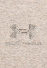 Lys beige bomuldsstof med en struktureret overflade. Broderet grå Under Armour-logo i midten, der fremhæver glatte detaljer og rene linjer.