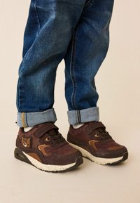 Brune sportssko med en tekstureret overflade, der har snørebånd og en velcrobånd. Udsmykket med et bjørnelogo og placeret på en spottet sål.