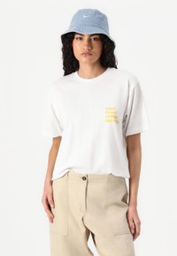 T-shirt bianca con scritta gialla "EASY PEASY LEMON SQUEEZY" sul lato sinistro del petto, abbinata a pantaloni beige a gamba larga e un cappello a secchiello blu chiaro.