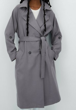 Personne portant un trench-coat gris ceinturé avec des boutons aux manches et aux poches, stylisé sur un top blanc.