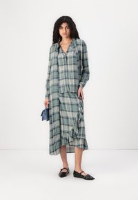 Grønn rutete skjorte og matchende skjørt, med volanger og en asymmetrisk skjorte. Modellen har på seg svarte leiligheter og holder en liten blå bag.