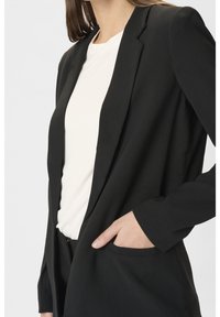 Personne portant un blazer noir sur une chemise blanche, une main reposant dans la poche du blazer sur un fond uni.