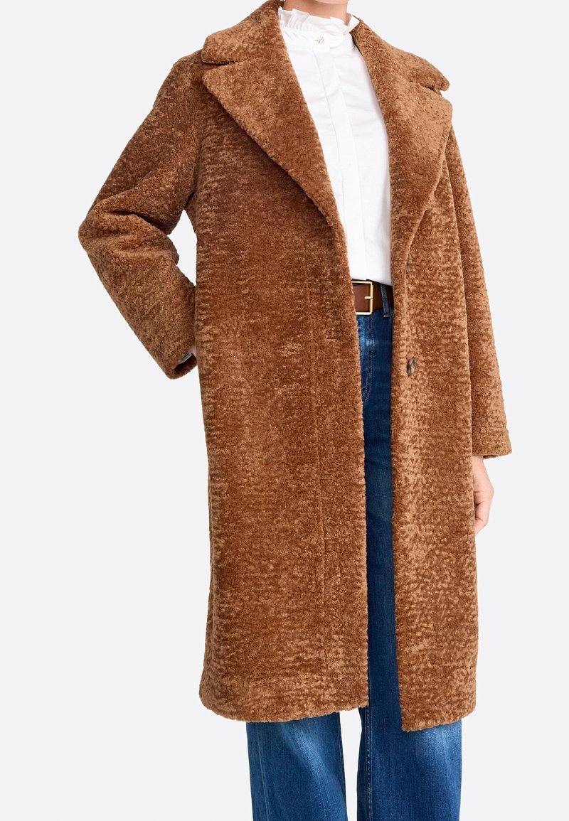 Cappotto in finta pelliccia marrone con ampio colletto, maniche lunghe e vestibilità morbida, chiusura con bottoni, abbinato a una camicia bianca e jeans blu.