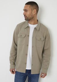 Světle hnědá button-up bunda s dvěma náprsními kapsami, vyrobena z bavlněné směsi, s uvolněným střihem, nošena přes bílé tričko a modré džíny.