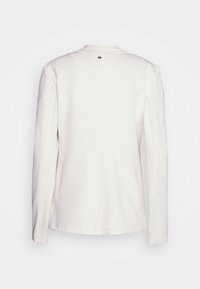 Bílá dlouhá bunda s dlouhým rukávem a malým kulatým kovovým emblémem na horní střední části zad, zobrazená na jednobarevném světlém pozadí.