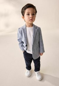 Ein Kind trägt einen hellgrauen Leinenblazer über einem weißen T-Shirt, blauen Hosen und weißen Sneakern. Der Blazer hat einen einzelnen Knopf und Taschen.