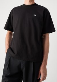 Czarny bawełniany t-shirt z okrągłym dekoltem, krótkimi rękawami i haftowanym logo na lewej piersi. Materiał ma gładką fakturę.