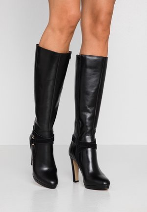 Bottes en cuir noires montant jusqu'aux genoux avec talons hauts et fermeture éclair latérale, portées sur des jambes nues, sur un fond uni.